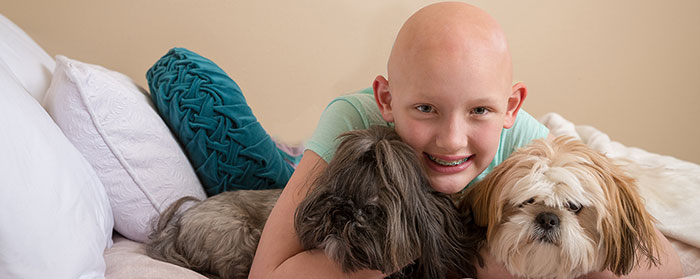 A girl with no hair smiling and hugging two cute dogs