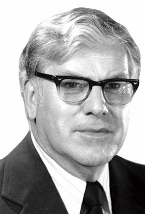 A black and white photo of a man wearing a black suit and black glasses