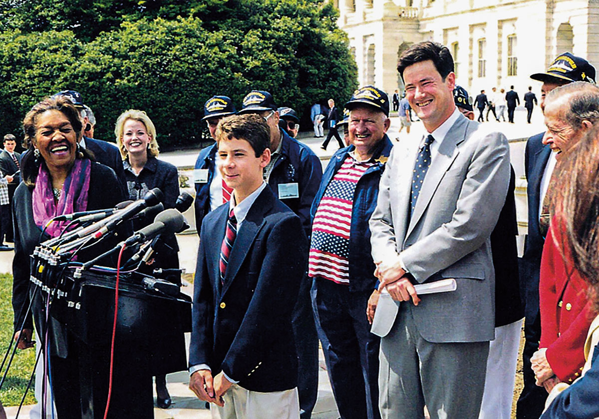 a young man in a suit speaking into a microphone with a crowd behind him