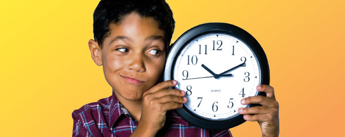 A boy wearing a plaid shirt looking at the round clock he is holding up next to his face